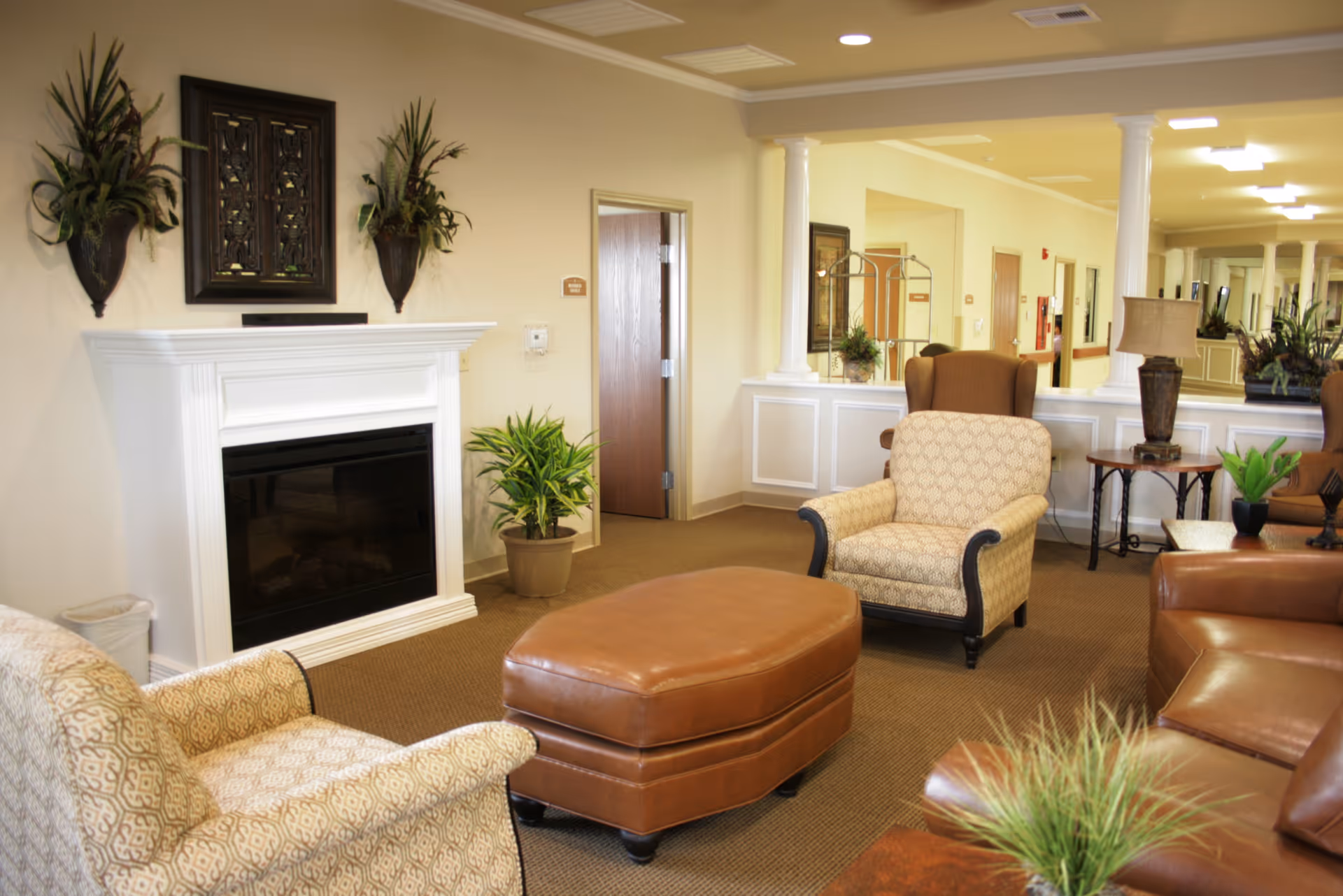 A comfortable senior living common room with upholstered armchairs, a leather ottoman, a white fireplace, and potted plants.