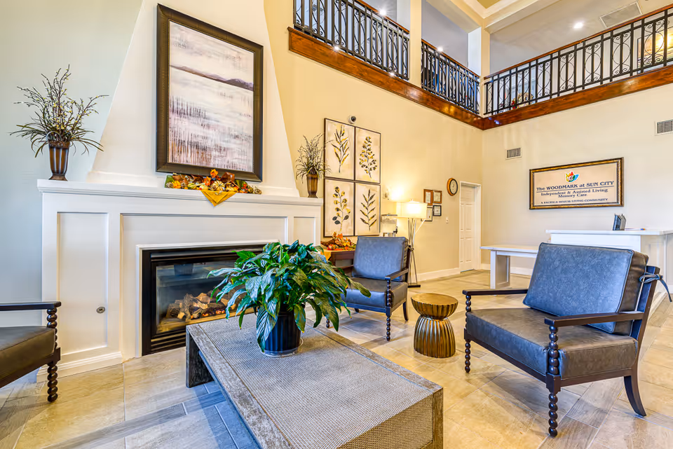 A bright and welcoming living room area in The Woodmark at Sun City featuring a fireplace with a framed painting above it, two dark wooden armchairs with gray cushions, a rectangular coffee table with a green potted plant, and decorative wall art. The room has a high ceiling with a balcony railing above and a reception desk in the background with a sign for The Woodmark at Sun City on the wall.