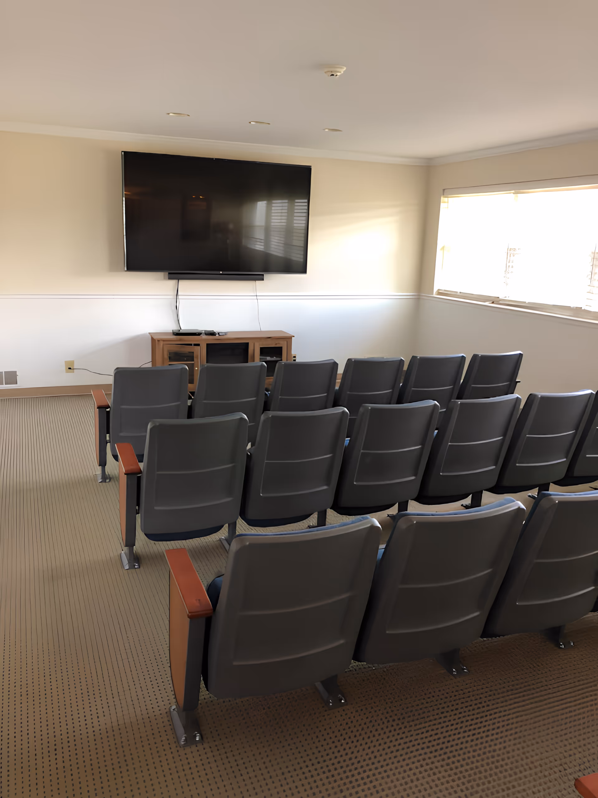 A seating area in a theater with gray chairs arranged in rows facing a large wall-mounted television, illuminated by natural light from a nearby window.