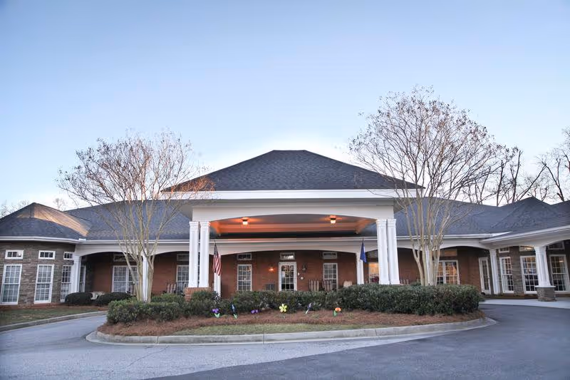 Front entrance of a senior living facility with a covered porte-cochère, white columns, brick facade, flags, and landscaped circular drive.
