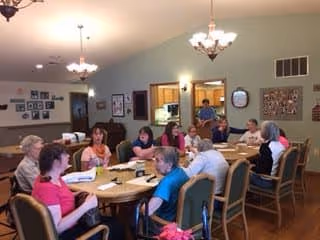 A group of elderly people sitting around a large rectangular table in a well-lit room, engaging in conversation or activities. The room has high ceilings with hanging light fixtures, framed pictures on the walls, and a person standing near a kitchen pass-through window in the background.