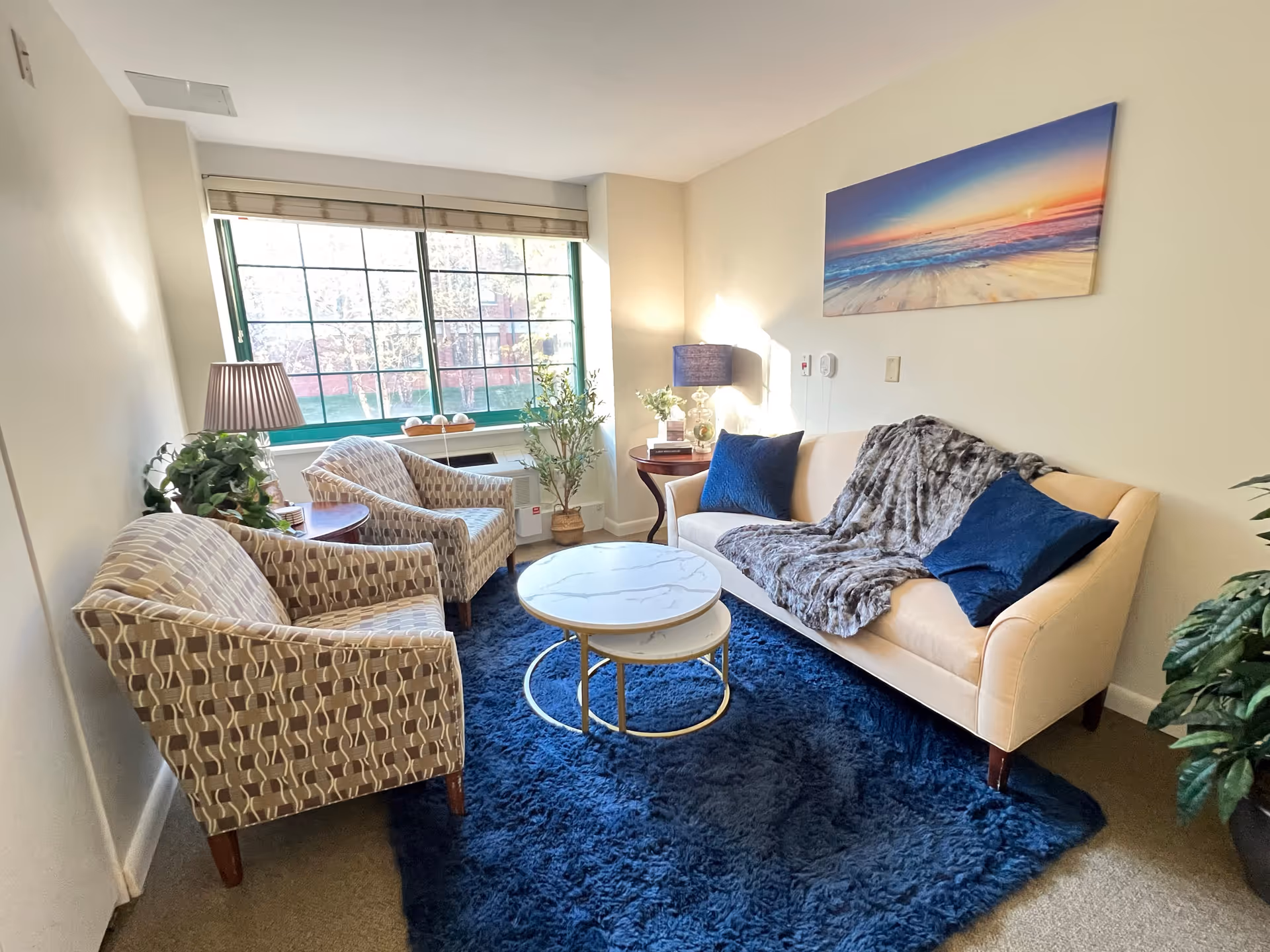 A cozy living room with a beige sofa adorned with two blue pillows and a gray throw blanket, two patterned armchairs, a round nested coffee table with a marble top, a blue shag rug, a side table with a lamp and decorative items, a large window with blinds letting in natural light, and a colorful beach sunset painting on the wall.