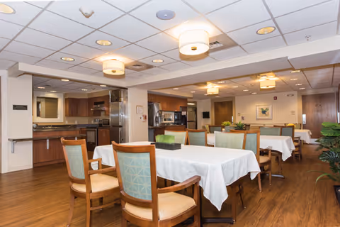 A spacious dining room in a senior living facility with several tables covered in white tablecloths and surrounded by wooden chairs with cushioned seats and backs. The room has wood flooring, recessed lighting, and ceiling-mounted light fixtures. In the background, there is a kitchen area with stainless steel appliances and wooden cabinetry. A framed picture hangs on the far wall, and there are some plants adding greenery to the space.