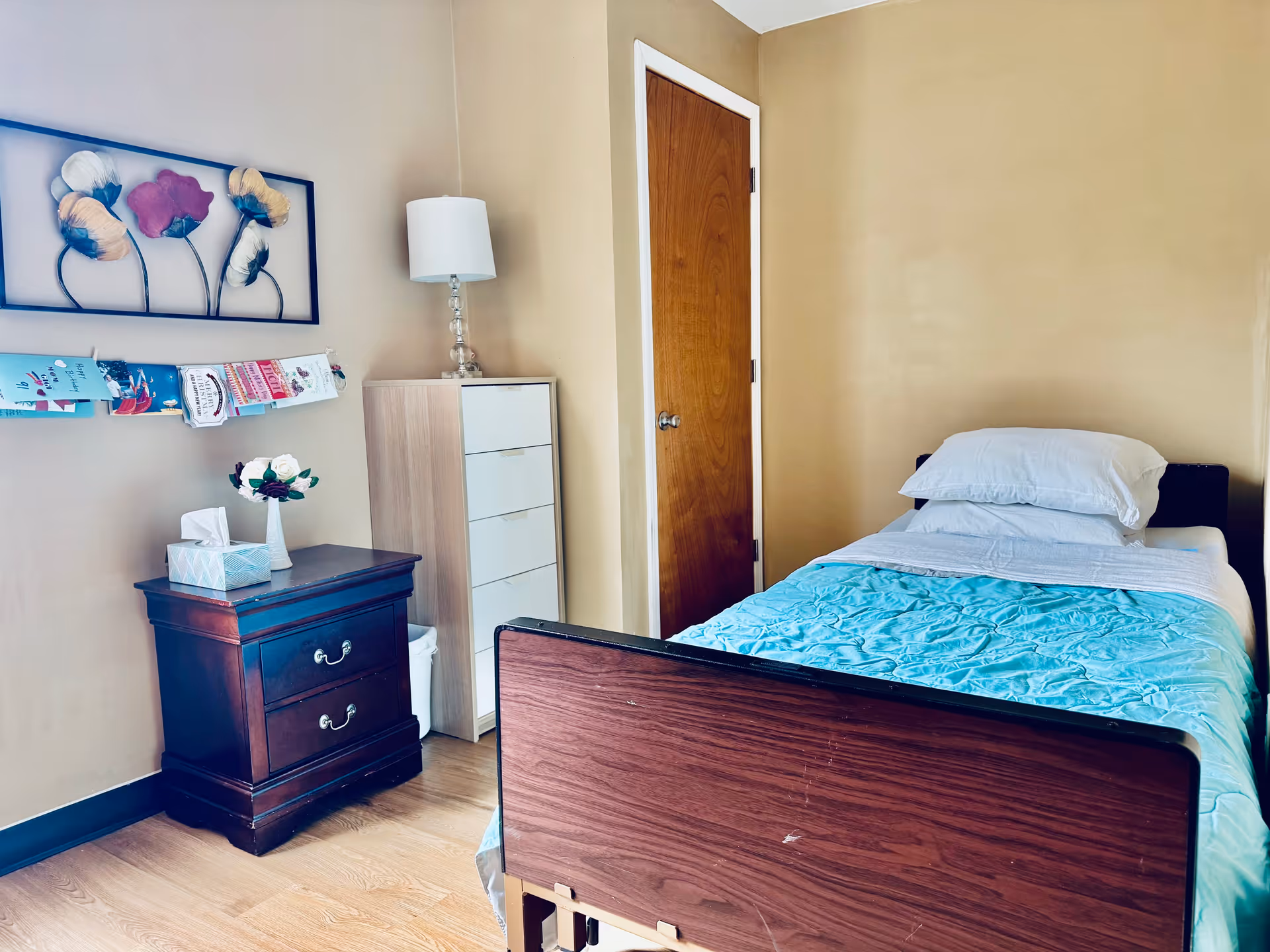 A small bedroom with a single bed made with white sheets and a light blue blanket. Next to the bed is a wooden nightstand with two drawers, a tissue box, and a small vase with flowers. Behind the nightstand is a tall white dresser with a lamp on top. On the wall above the nightstand is a decorative metal wall art with flowers and a string holding several cards or papers. The room has beige walls and a wooden door.