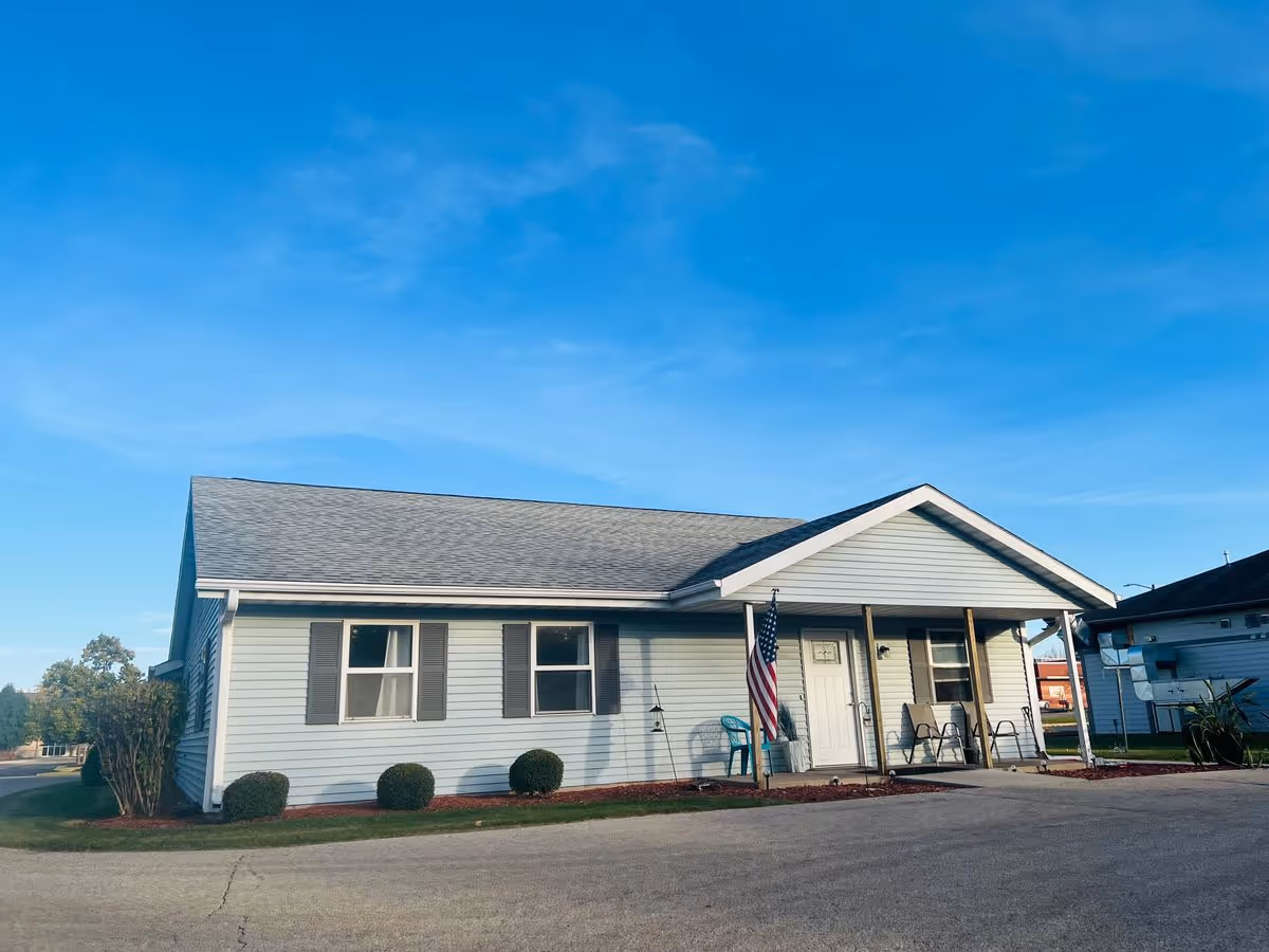 Single-story light-blue building with a covered front porch, an American flag, and chairs under a clear blue sky.
