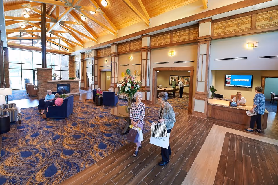 Spacious senior living facility lobby with high wooden beam ceiling, large windows, and a fireplace. Several elderly people are seated on blue chairs and sofas, while two women carrying shopping bags are walking and talking near a round table with a flower arrangement. A reception desk with a staff member and a visitor is visible on the right side, and a game room with a pool table is seen in the background.