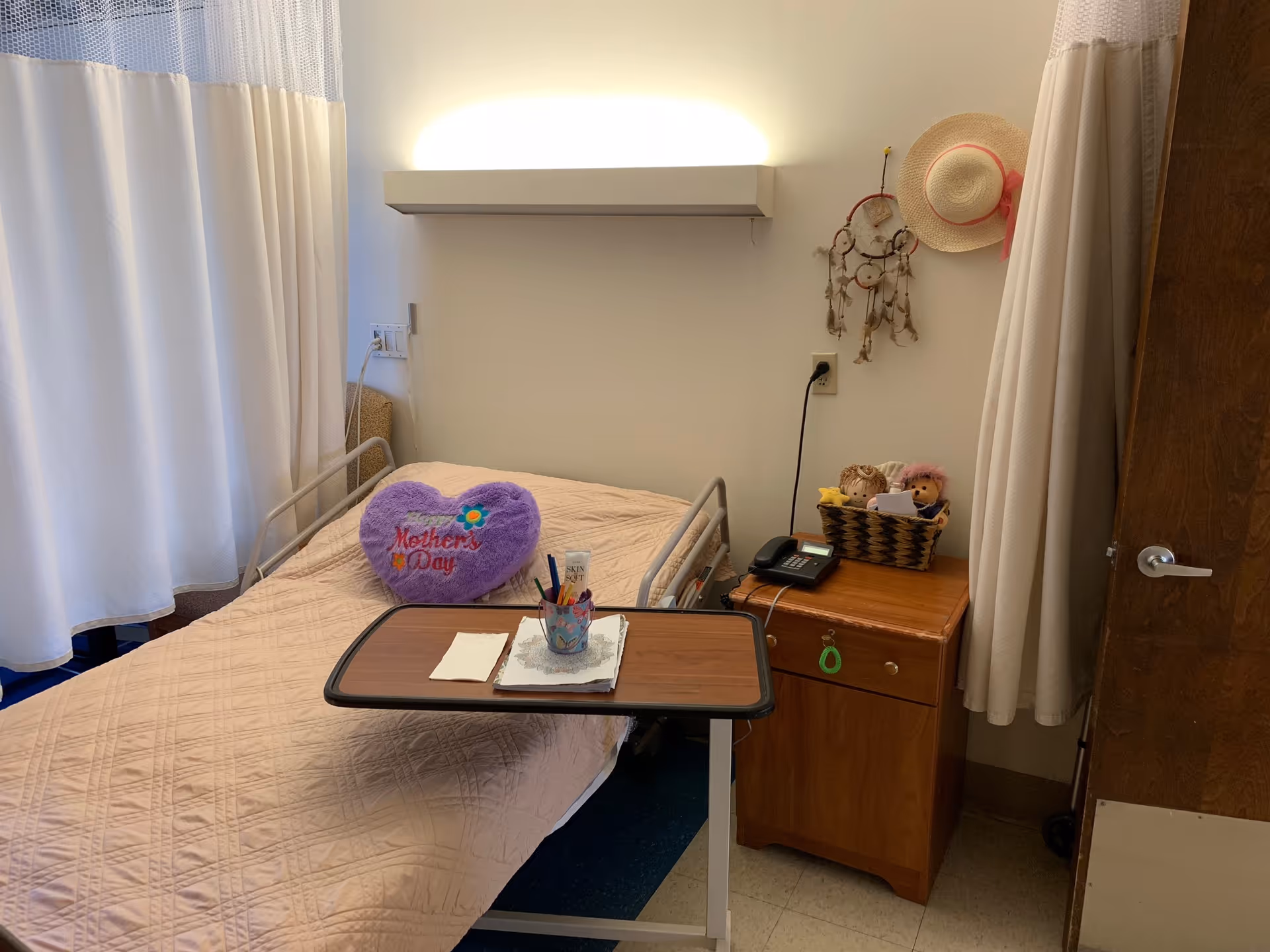 A small bedroom in a senior living facility with a single bed covered by a beige quilt. On the bed is a purple heart-shaped pillow with the words 'Mother's Day' embroidered on it. A wooden bedside table holds a telephone and a basket with two stuffed dolls. A dreamcatcher and a straw hat hang on the wall above the table. A wooden overbed table with pens and paper is positioned over the bed. White curtains partially enclose the bed area.