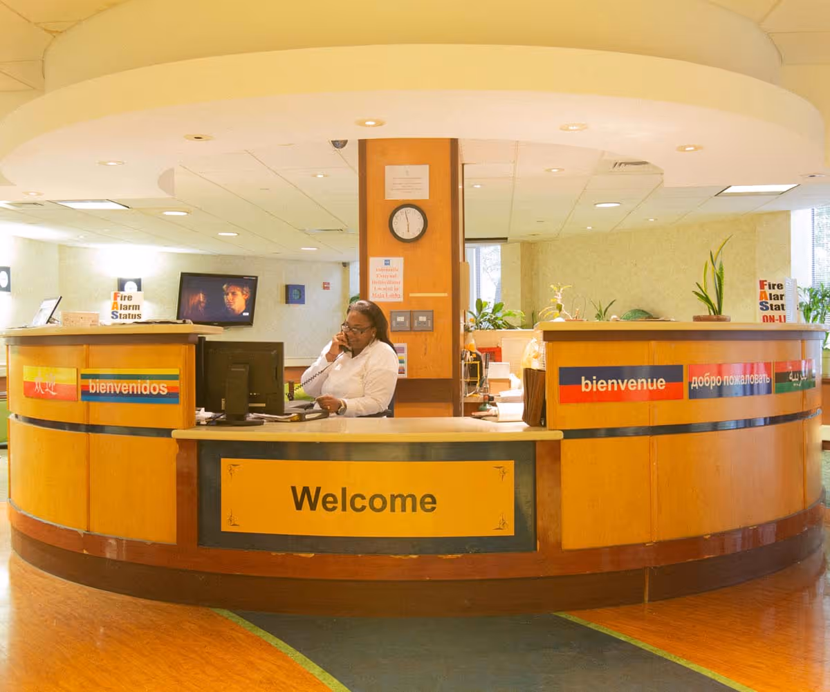Reception desk area at Downtown Brooklyn Nursing and Rehabilitation with a staff member talking on the phone. The desk has multilingual welcome signs including 'bienvenidos' and 'bienvenue'. There is a clock on the wall behind the desk and a TV screen mounted on the wall.