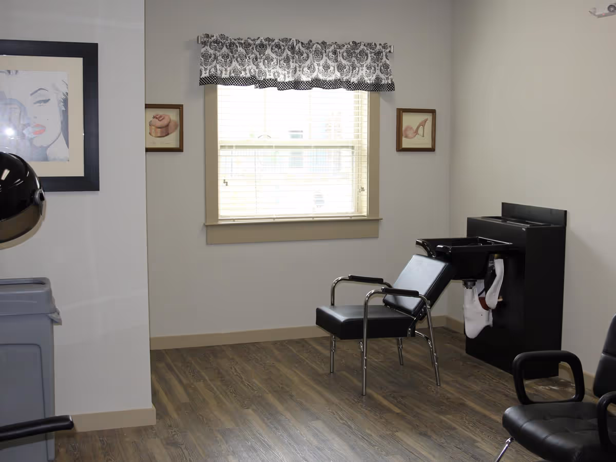 A small hair salon area with a black salon chair in front of a black hair washing station. The room has wood flooring, a window with white blinds and a black and white patterned valance, and framed artwork on the walls. There is also a black salon dryer hood partially visible on the left side.