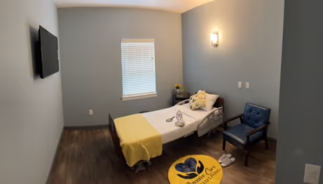 A small, simple bedroom with a single bed covered with white sheets and a yellow blanket. There is a blue cushioned chair next to the bed, a small table with a flower vase, and a window with blinds. A flat-screen TV is mounted on the wall opposite the bed. The floor has a round yellow rug with the Precisive Care I and II logo. The walls are painted light gray.