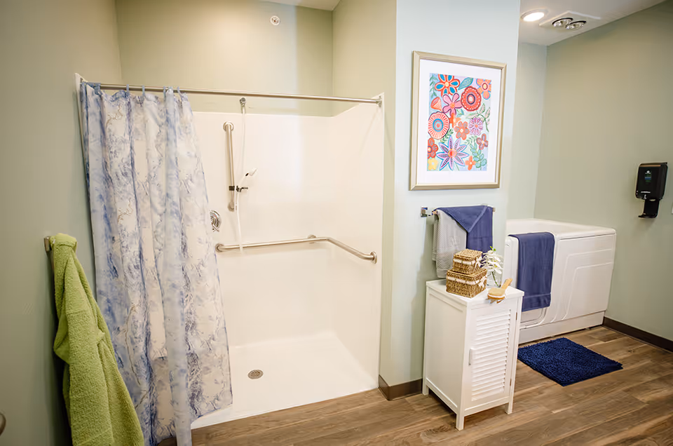 A bathroom area with a walk-in shower featuring a blue and white patterned shower curtain and grab bars. There is a green towel hanging on the wall to the left. To the right of the shower, there is a small white cabinet with woven baskets and a small plant on top. Above the cabinet, a colorful floral artwork is framed on the wall. Further right, there is a white washing machine with a blue towel draped over it and a dark blue bath mat on the wooden floor. A soap dispenser is mounted on the wall above the washing machine.