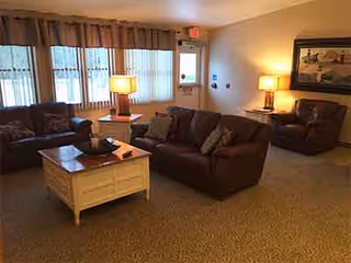 A cozy living room area with brown leather sofas and armchairs arranged around a white wooden coffee table. The room is softly lit by two table lamps, one on a side table and one on a small cabinet. Large windows with vertical blinds allow natural light to enter the space. A framed painting hangs on the wall above one of the armchairs.