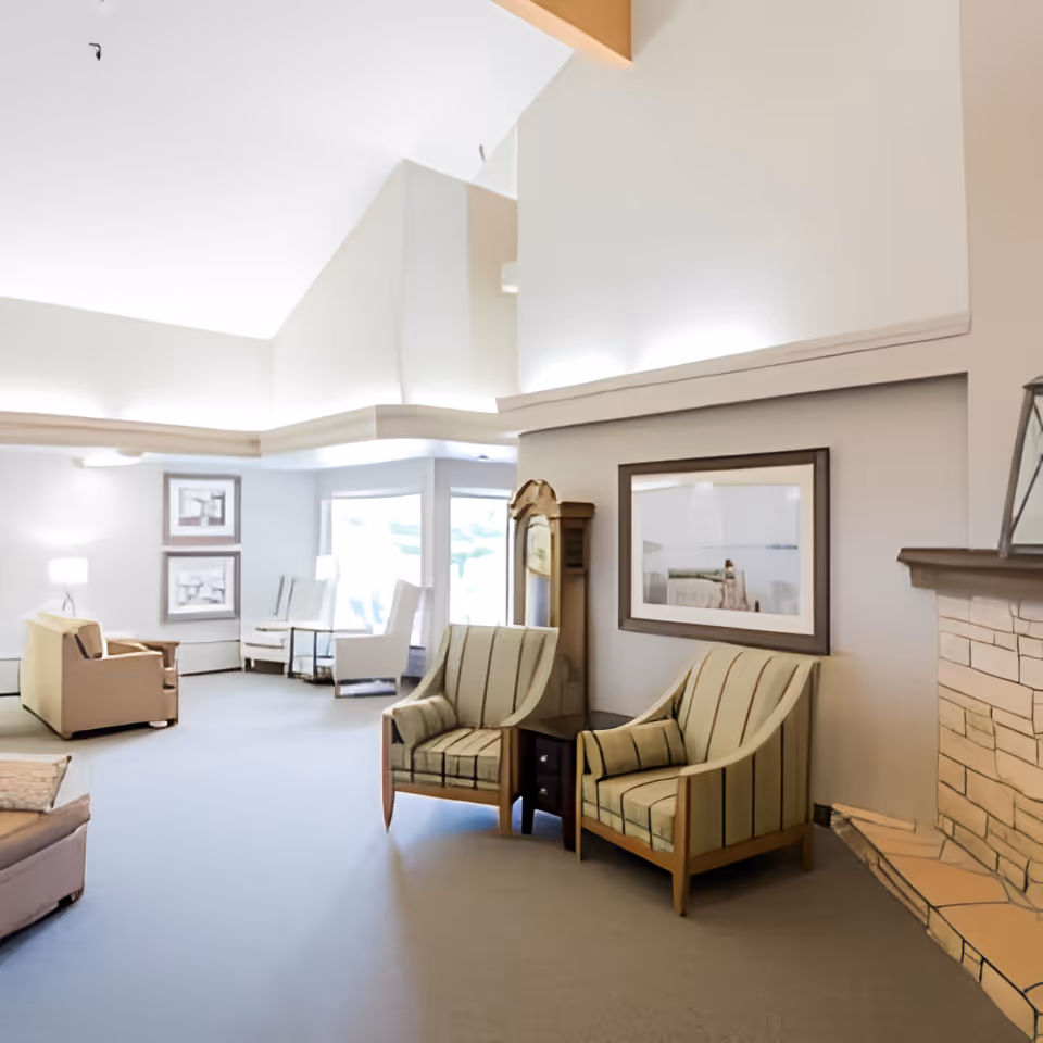 A bright and spacious living room area with high ceilings, featuring two striped armchairs next to a small wooden table, a stone fireplace on the right, and additional seating including armchairs and a sofa in the background near large windows letting in natural light. The walls are decorated with framed artwork.