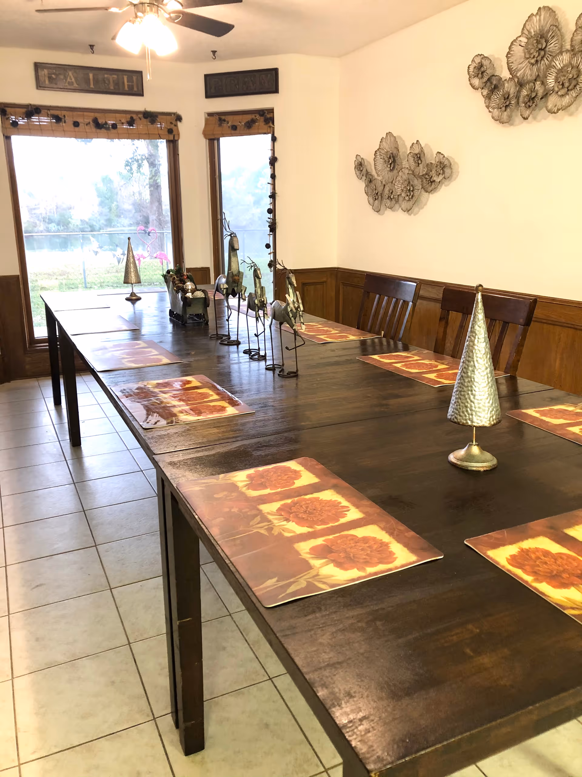 A long wooden dining table set with floral placemats and decorative metal sculptures in the center. The room has tiled flooring, wooden chairs, and large windows with wooden blinds. Wall decorations include metal flower art and framed signs reading 'FAITH' and 'PRAY'.