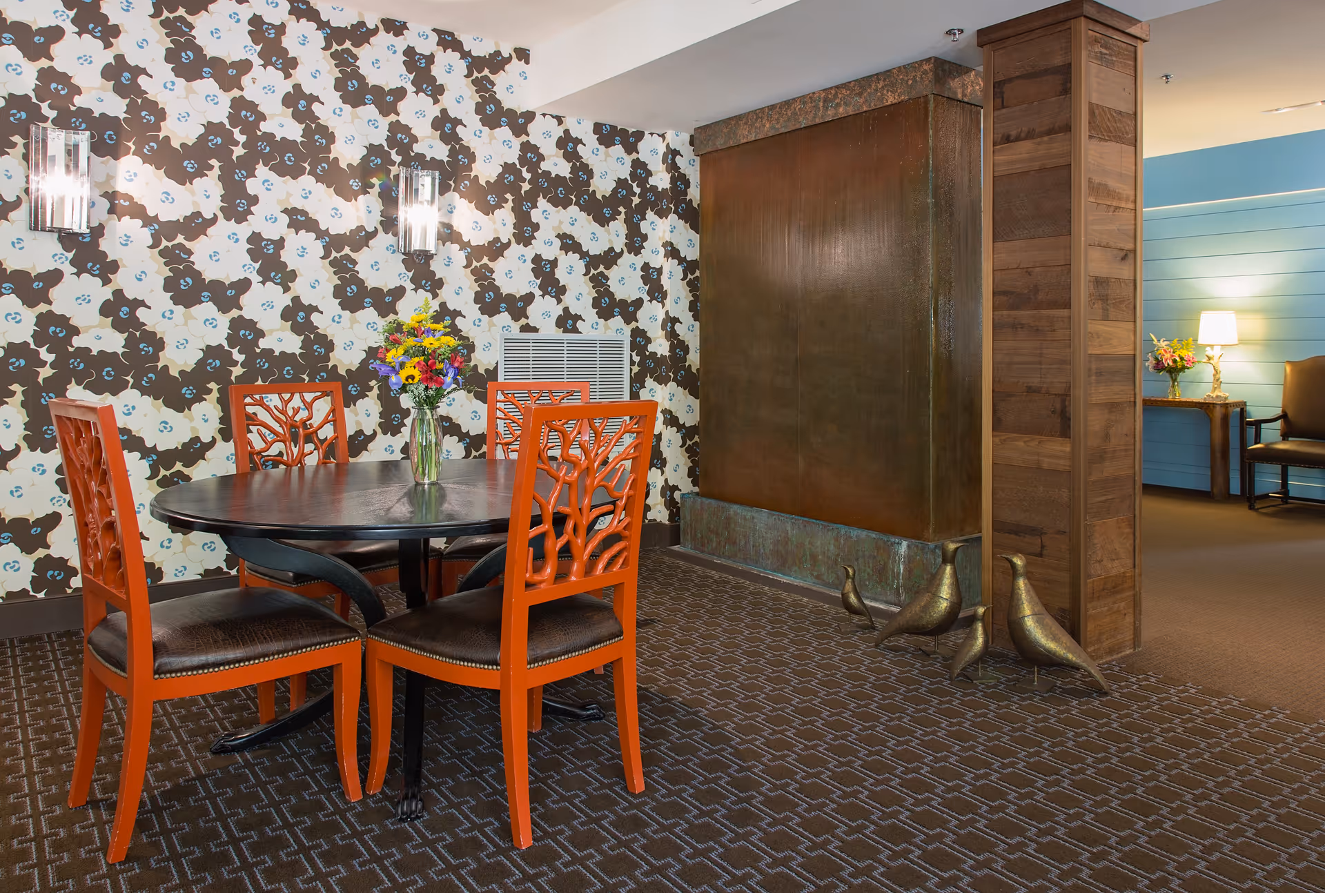 A cozy interior space featuring a round black table with four orange chairs that have a tree branch design on the backrests. A vase with colorful flowers is placed on the table. The wall behind the table has a floral patterned wallpaper in brown, white, and blue. To the right, there is a large bronze water feature with three bronze bird sculptures at its base. In the background, a small table with a lamp and another vase of flowers is visible against a blue shiplap wall.