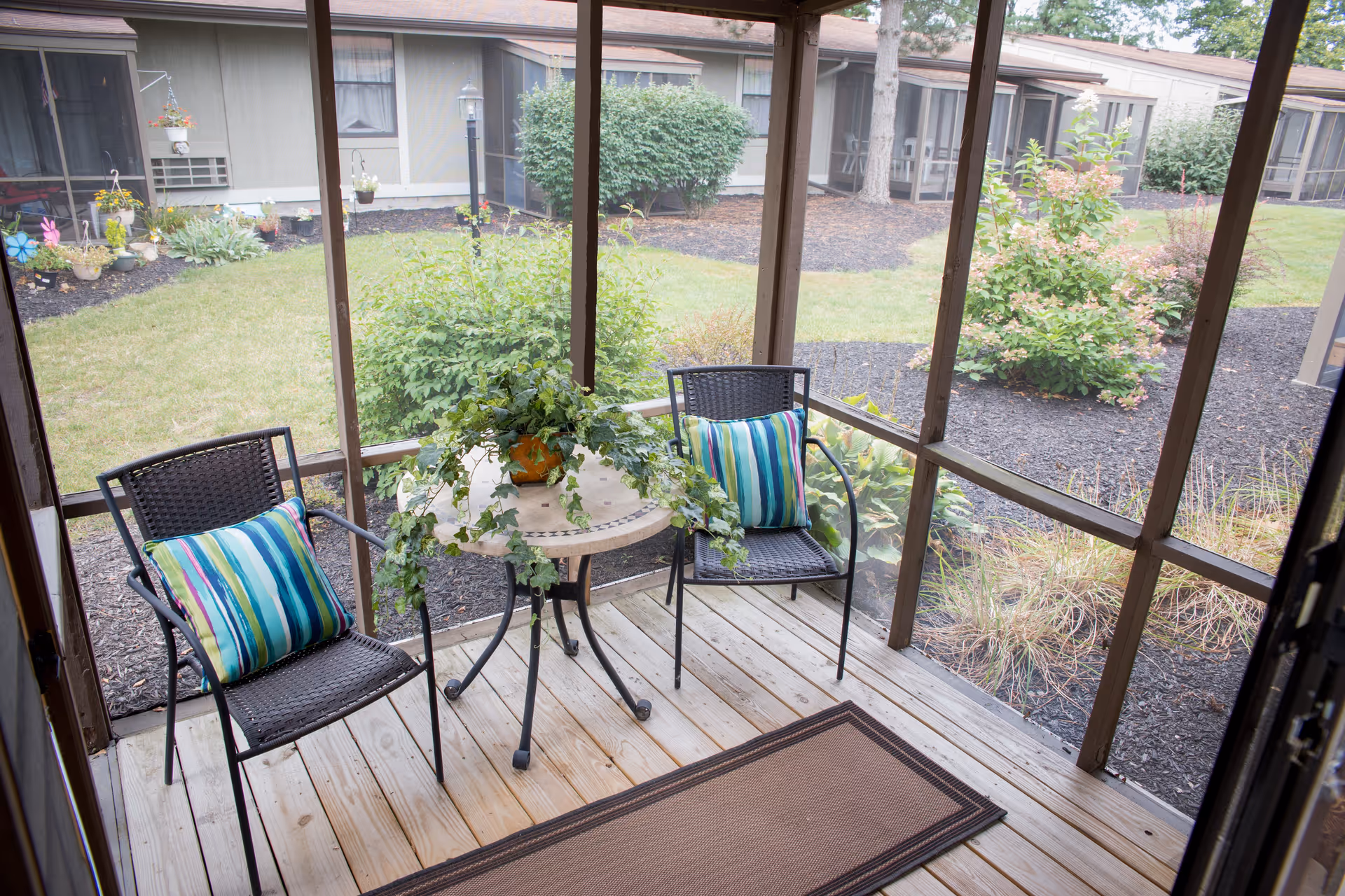 A screened-in porch with two black wicker chairs each with a colorful striped cushion, a small round table with a potted green plant, and a brown floor mat. Outside the porch, there is a grassy courtyard with bushes and a building with multiple screened windows and doors.