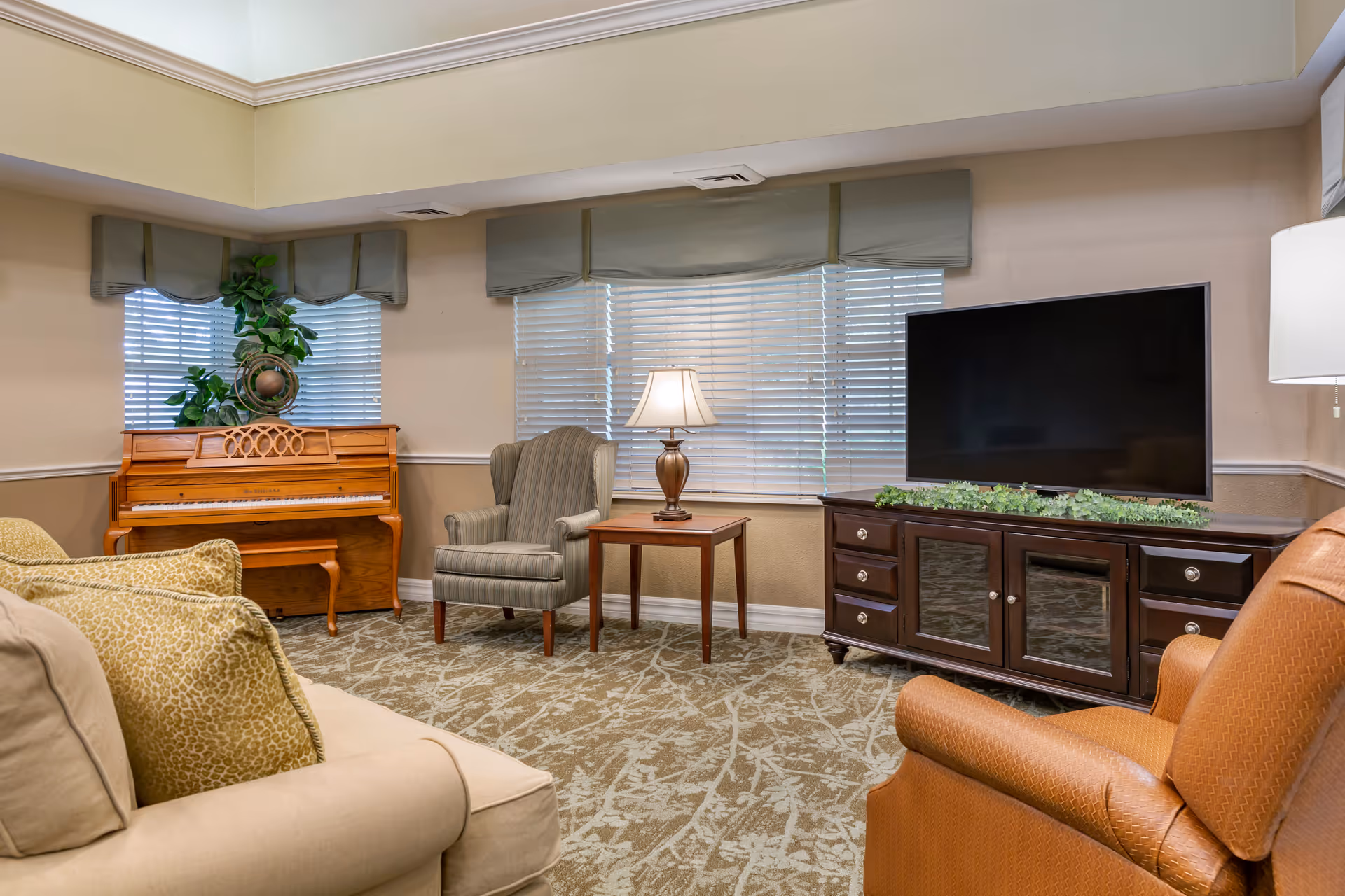 A cozy living room with a beige sofa, a striped armchair, and an orange armchair arranged around a wooden TV stand with a large flat-screen TV. There is a wooden piano with a plant on top near the windows with blinds and valances. A table lamp sits on a small wooden side table between the armchair and the TV stand. The room has a patterned carpet and soft, neutral-colored walls.