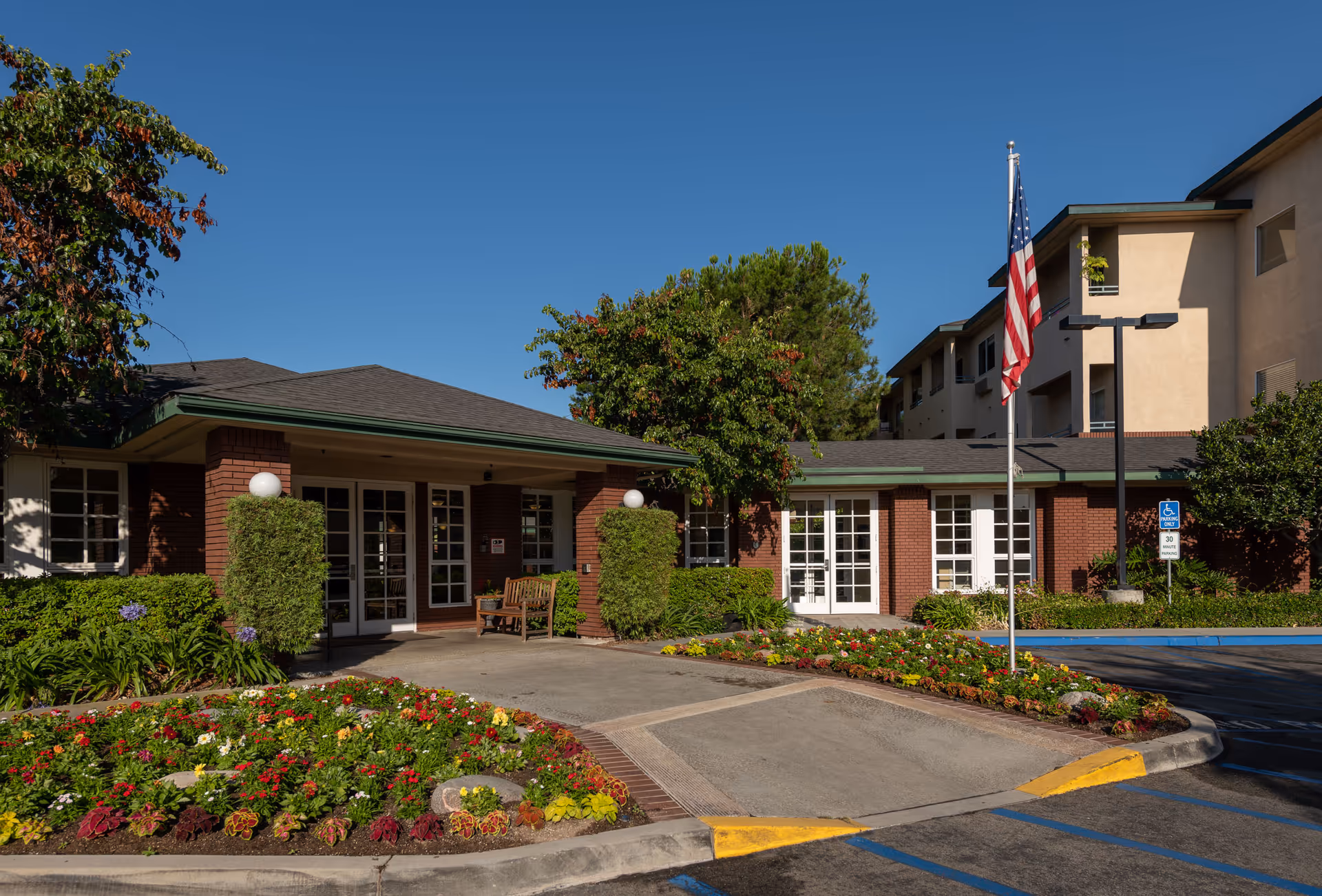 Exterior view of Carmel Village Retirement Community showing a brick building with large windows and a covered entrance. There are well-maintained flower beds with colorful flowers and shrubs in front, an American flag on a flagpole, and a clear blue sky above.