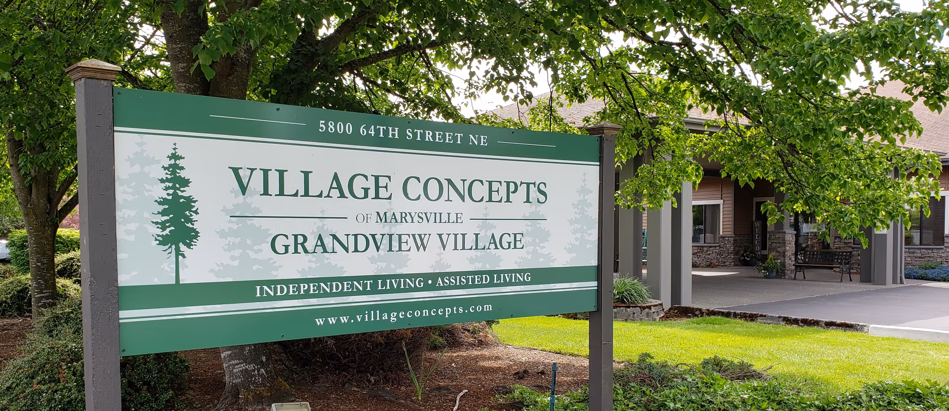 Outdoor view of a large green and white sign for Village Concepts of Marysville - Grandview Village, an independent and assisted living facility, located at 5800 64th Street NE. The sign is surrounded by trees and landscaping, with part of the building entrance visible in the background.