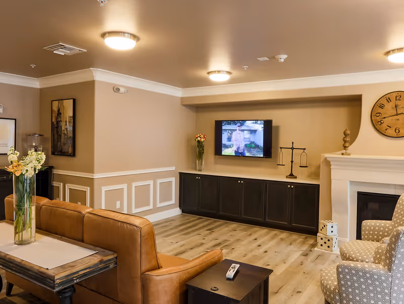 A cozy living room area with a brown leather sofa, a patterned armchair, a wooden coffee table with a vase of flowers, a wall-mounted TV, a fireplace with decorative items, and a large wall clock. The room has beige walls, wood flooring, and ceiling lights.