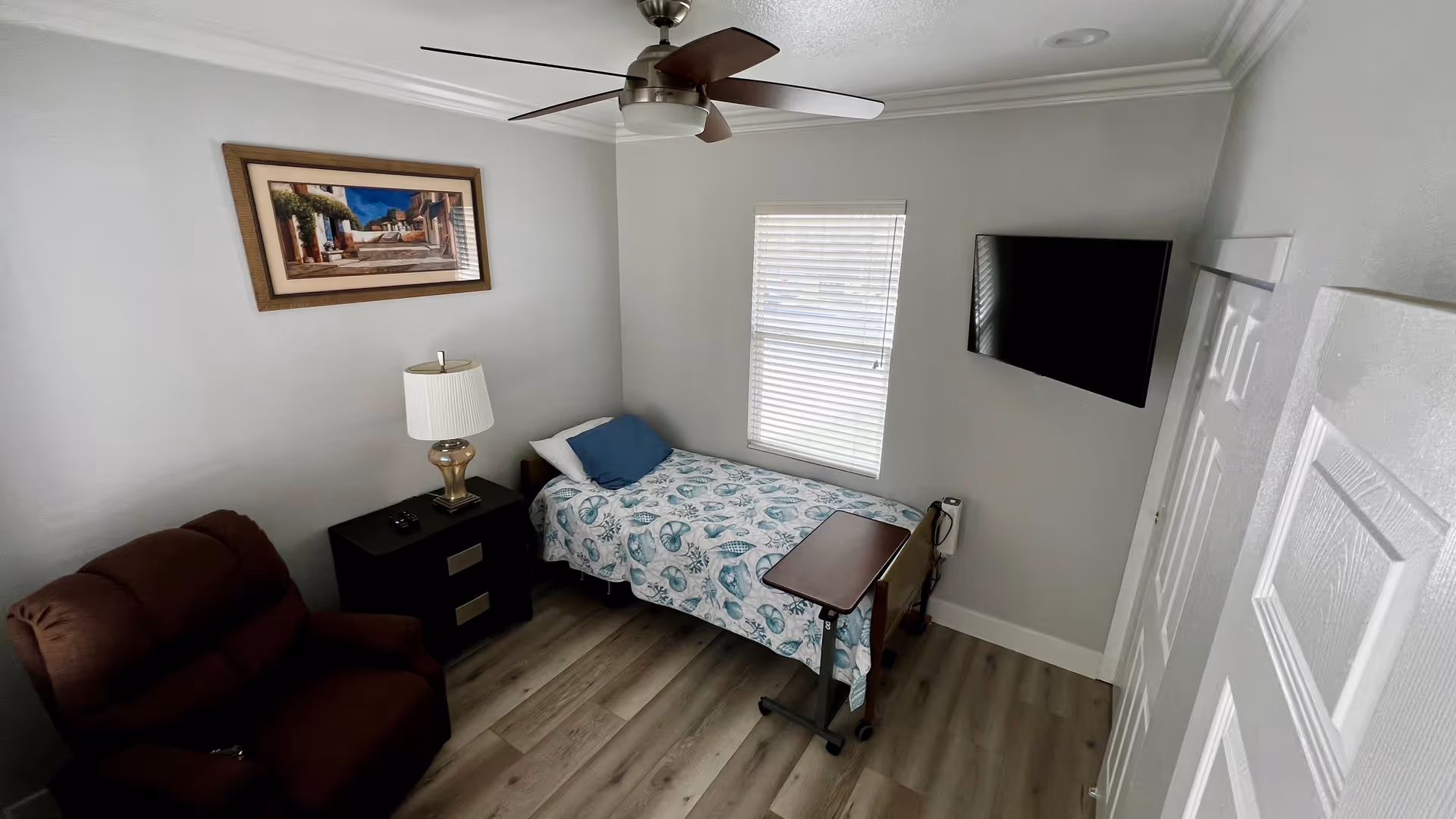 A small senior care bedroom with a single bed covered in a seashell patterned bedspread, a blue pillow, a dark brown recliner chair, a black nightstand with a lamp and remote controls, a ceiling fan, a window with blinds, a wall-mounted flat screen TV, and a framed painting on the wall.