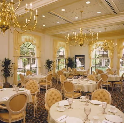 Elegant dining room with round tables covered in white tablecloths, set with plates, cups, and glasses. The room features beige upholstered chairs, large windows with yellow curtains, gold chandeliers hanging from the ceiling, and potted plants near the windows.