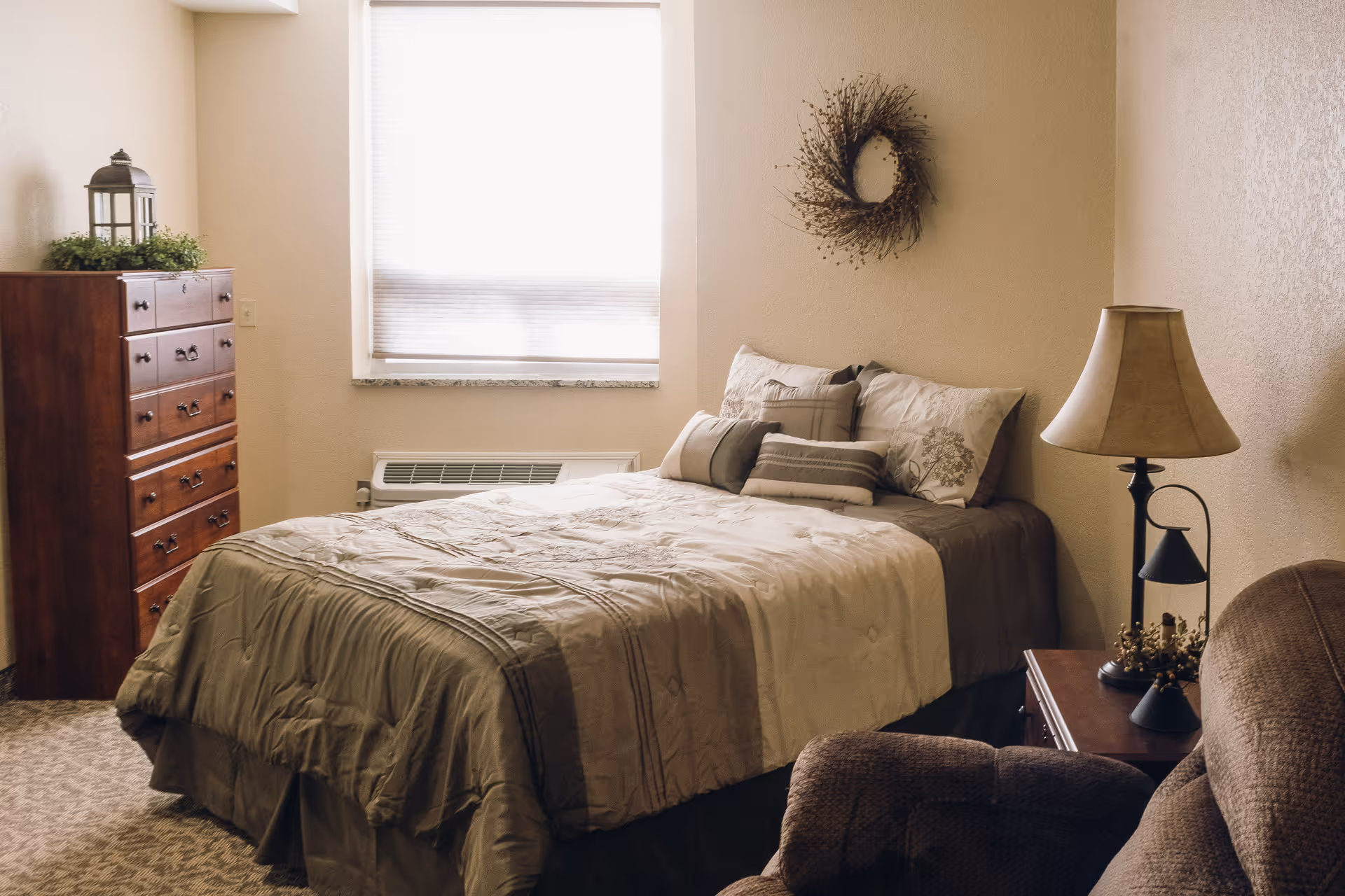 A cozy bedroom with a made bed, decorative pillows, a wooden dresser by the window, a bedside table with a lamp, and an armchair.