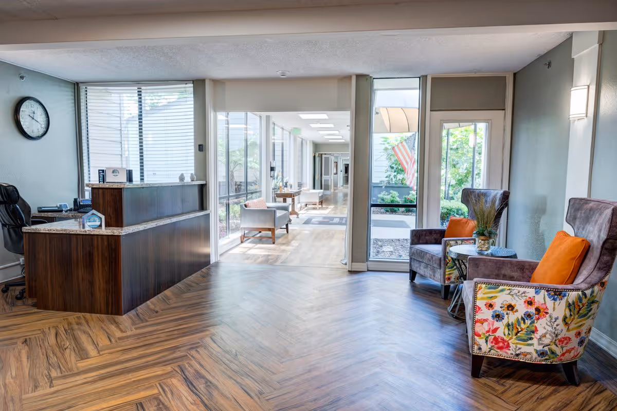 Reception area of Greenhaven Place with a wooden front desk on the left, a black office chair behind it, and a clock on the wall. To the right, there are two armchairs with floral upholstery on the sides and orange cushions, separated by a small round table with a plant. Large windows and glass doors let in natural light, showing a hallway and outdoor greenery.