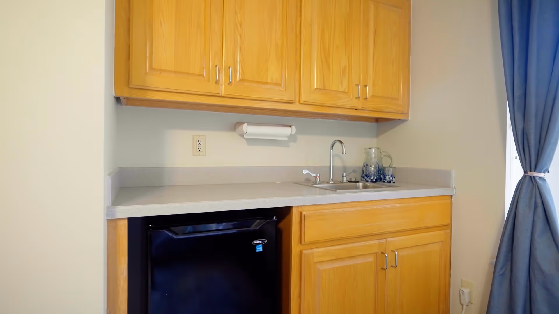A small kitchenette area with light wooden cabinets above and below a gray countertop. There is a small black mini-fridge under the counter, a stainless steel sink with a faucet, a paper towel holder mounted on the wall, and a glass pitcher with a matching glass on the counter. A blue curtain is partially visible on the right side of the image.