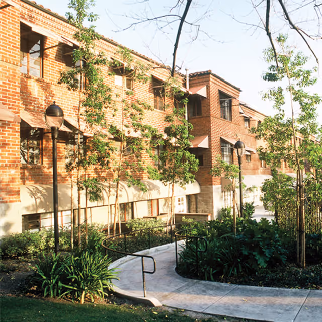 Exterior view of a brick building with multiple windows and awnings, surrounded by young trees and landscaped greenery. A curved concrete pathway with handrails leads through the garden area in front of the building.