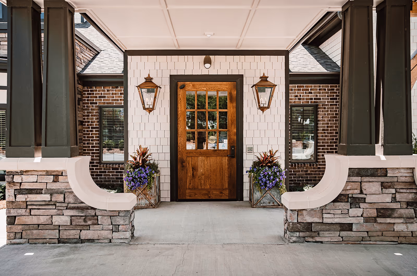 Front entrance of a building with a wooden door featuring glass panes, flanked by two large lantern-style wall lights and two planters with purple flowers and greenery. The entrance is framed by stone pillars and white siding with brick walls on either side.