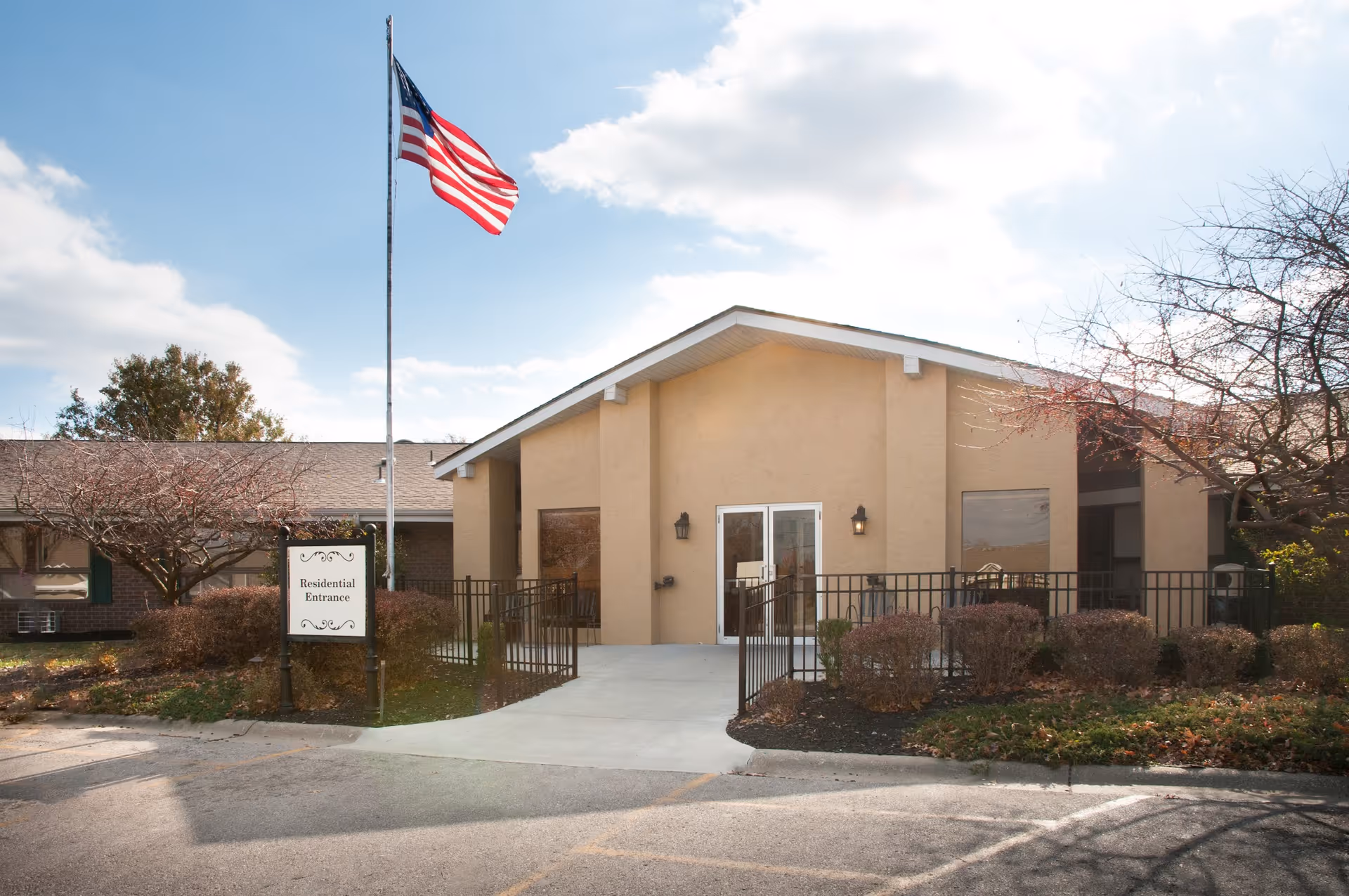 Exterior view of the Life Care Center of Osawatomie showing the residential entrance with a beige building, a concrete pathway, black metal railing, bushes, and an American flag on a flagpole against a partly cloudy sky.