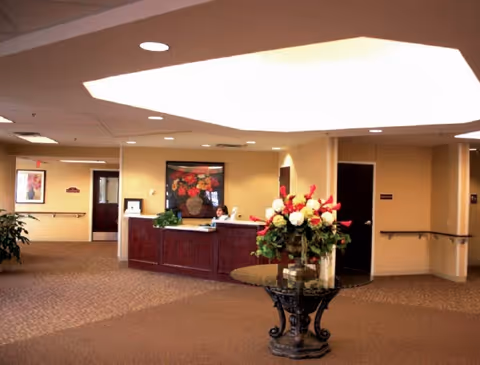 Interior view of a senior living facility reception area with a wooden front desk, a receptionist seated behind it, a round glass table with a large floral arrangement in the foreground, beige walls, carpeted floor, and framed artwork on the walls.