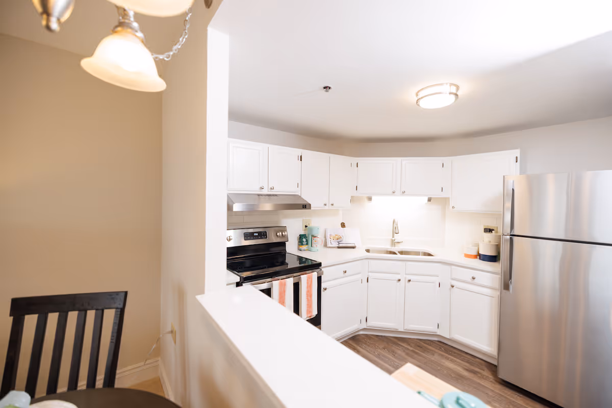 Bright kitchen with white cabinets, stainless steel refrigerator and stove, double sink, and wooden flooring. A partial view of a dining table with a black chair is visible on the left side.
