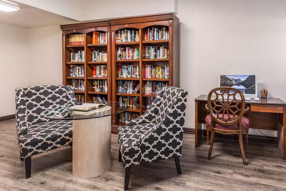 A cozy reading and computer area in a senior living facility featuring two patterned armchairs around a small glass-top table with an open book, a large wooden bookshelf filled with books, and a wooden desk with a computer and a wooden chair with a cushioned seat.