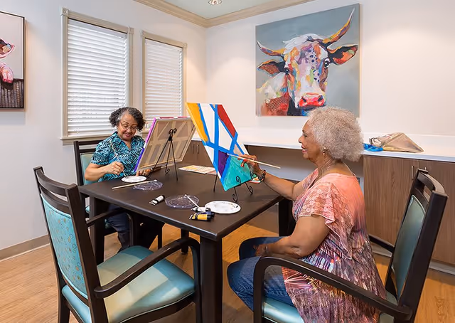 Two elderly women sitting at a table in a well-lit room, painting on canvases placed on easels. One woman is wearing a blue patterned shirt and glasses, while the other is wearing a pink patterned blouse. The room has light-colored walls, two windows with blinds, and a colorful painting of a cow on the wall behind them.