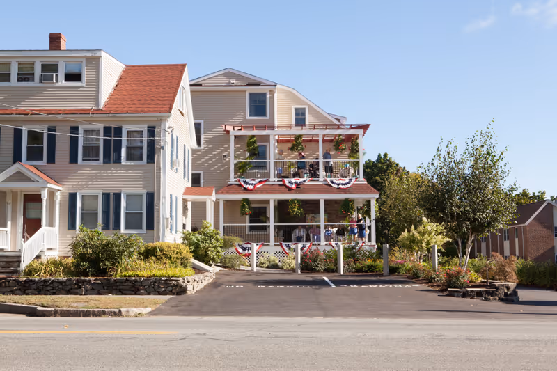 Exterior view of a multi-story beige building with red roof shingles and white trim, decorated with red, white, and blue bunting on the balconies. The building is surrounded by greenery and a paved parking area in front under a clear blue sky.