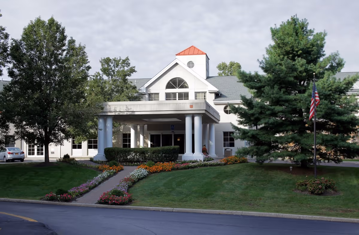 Front entrance of a nursing and rehabilitation center with a covered portico, white columns, landscaped flowerbeds and an American flag.