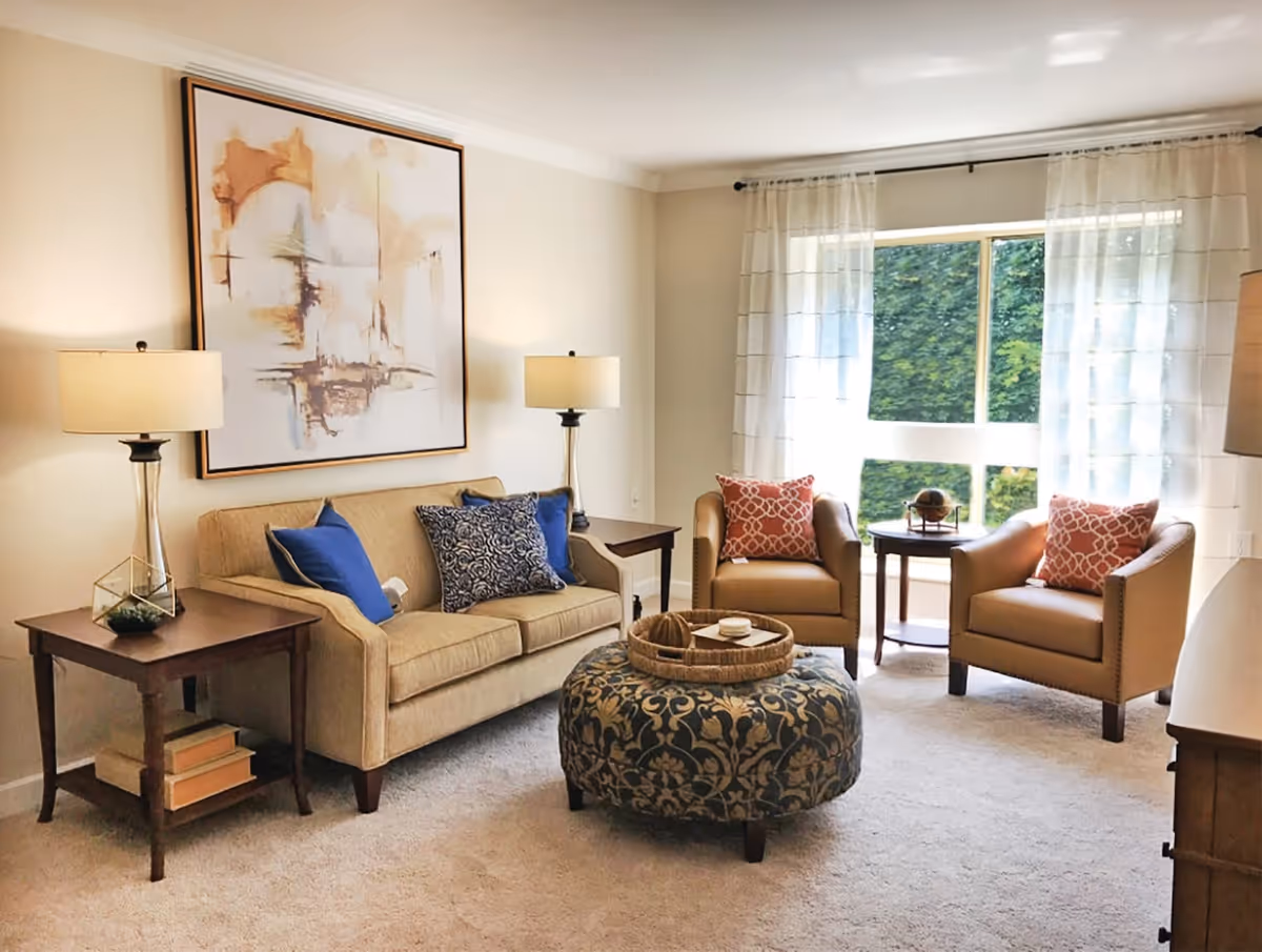 A cozy living room with a beige sofa adorned with blue and patterned cushions, two tan armchairs with orange patterned pillows, a round patterned ottoman with a wicker tray on top, two wooden side tables each with a lamp, a large abstract painting on the wall, and a window with sheer white curtains letting in natural light.