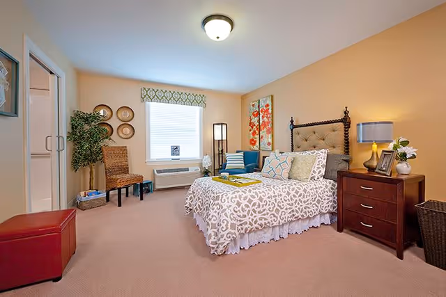 A cozy bedroom with a patterned bedspread on a bed with a tufted headboard, a wooden nightstand with a lamp and flowers, a blue armchair with pillows, a window with a decorative valance, a wicker chair, and a potted plant. The walls are painted a warm beige color.