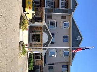Exterior front view of a multi-story senior living facility building with beige and gray siding, multiple windows, a stone entrance, and an American flag on a flagpole in front. A yellow car is parked near the entrance.