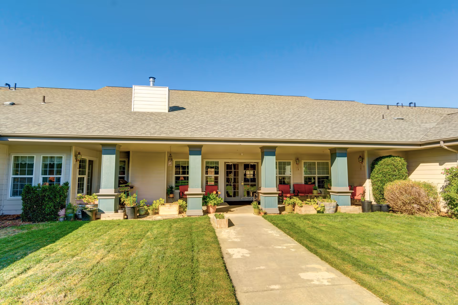 Exterior view of a single-story senior living facility building with a covered porch supported by green columns. The porch has red cushioned seating and various potted plants. A concrete walkway leads through a well-maintained green lawn to the entrance. The sky is clear and blue.