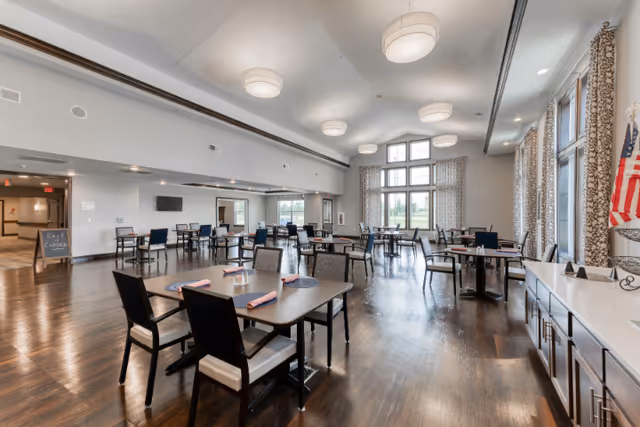A spacious dining room with multiple tables and chairs arranged neatly. The room features large windows with patterned curtains allowing natural light to fill the space. The floor is wooden, and the ceiling has several round light fixtures. An American flag is visible on the right side near a counter with cabinets underneath.
