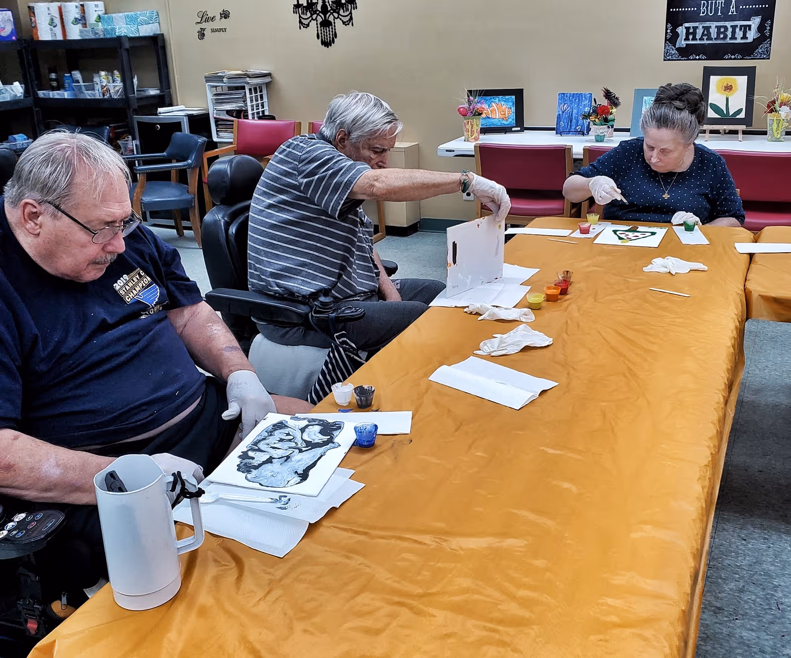 Three elderly individuals seated at a long table covered with a yellow tablecloth, engaged in painting activities. Each person has art supplies such as paint cups and brushes in front of them. The room has shelves with various items and artwork displayed on a table and walls in the background.