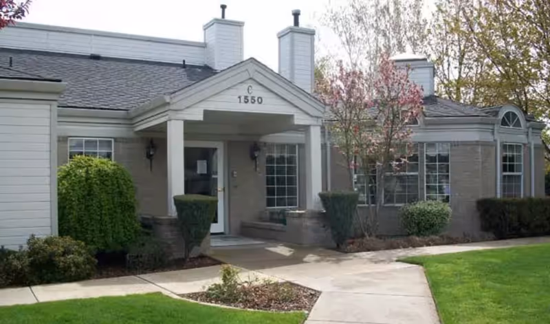 Exterior view of a single-story building with a covered entrance, labeled with the number 1550 above the door. The building is surrounded by well-maintained landscaping including green grass, bushes, and trees with some pink blossoms.