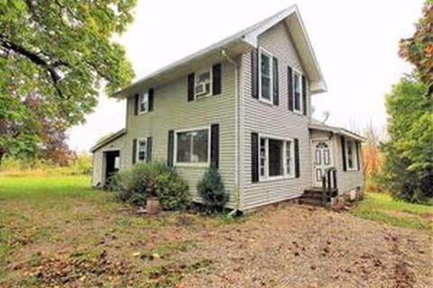 A two-story white house with siding sits in a grassy yard with trees.