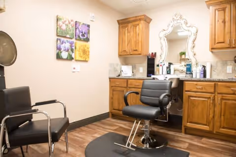 Salon-style interior with a black styling chair in front of an ornate mirror, wooden cabinets, and a second waiting chair on wood flooring.