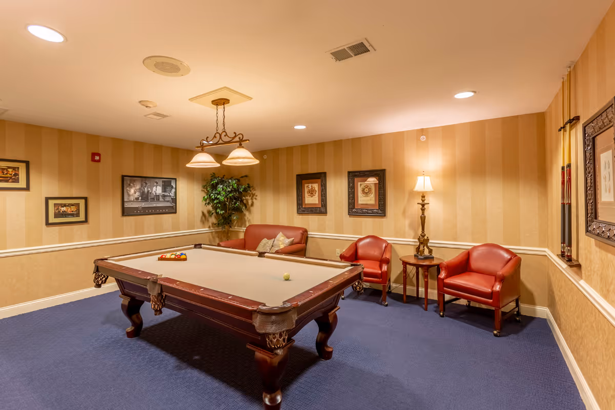 A cozy game room with a pool table in the center, a brown leather loveseat, two red leather armchairs, a wooden side table with a lamp, framed pictures on the beige striped walls, and a potted plant in the corner.