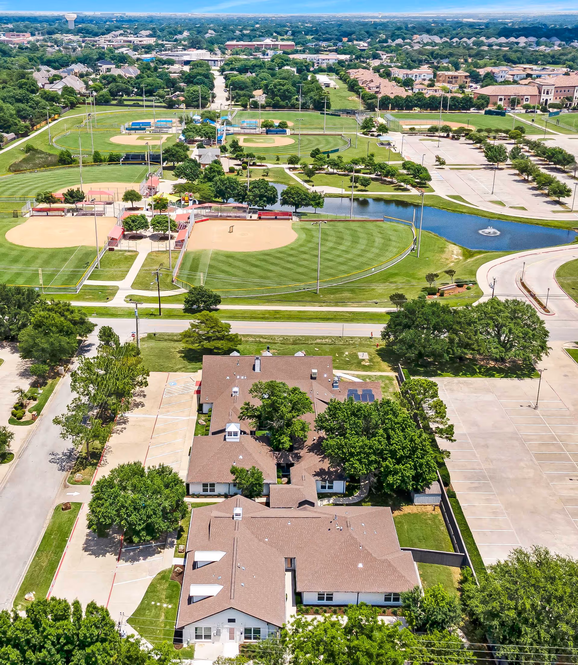 Aerial view showing a senior living facility complex surrounded by trees, parking lots, a pond, and several baseball fields.
