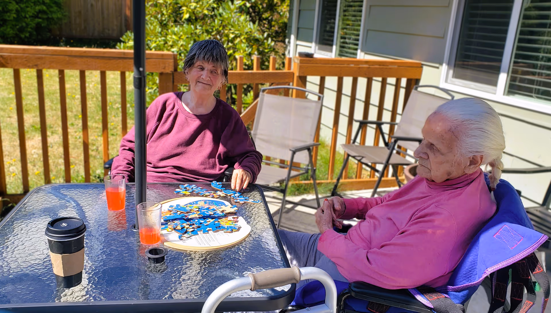 Two elderly women sitting outside on a patio at a glass table with a puzzle and drinks. One woman is wearing a maroon sweater and the other a pink sweater, both seated in wheelchairs. The patio has wooden railings and some chairs in the background.